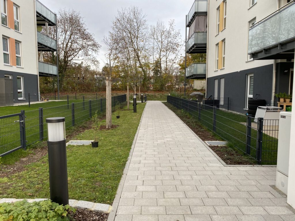 gepflasterter Erschließungsweg zwischen den Gebäuden, begleitend von Rasen mit Baumpflanzungen und Beleuchtung